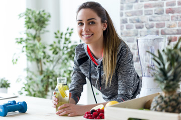 Sporty young woman looking at camera while drinking lemon juice in the kitchen at home.