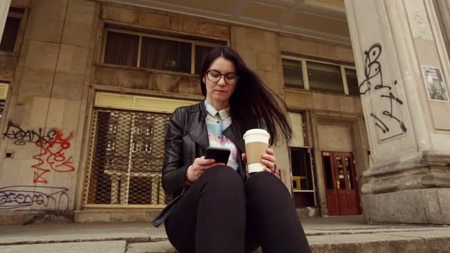 Charming girl in the city center, in a black leather jacket and glasses, browsing social networks, news, on your smartphone