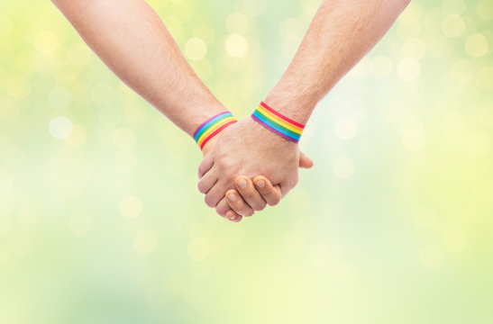 lgbt, same-sex relationships and homosexual concept - close up of male couple wearing gay pride rainbow awareness wristbands holding hands over green lights background