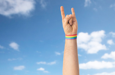 lgbt, same-sex relationships and homosexual concept - close up of male hand wearing gay pride awareness wristband showing rock or hand-horns sign over blue sky and clouds background