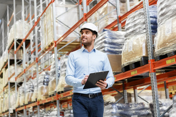 logistic business, shipment and people concept - businessman in helmet with clipboard checking goods at warehouse