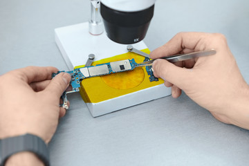 Repairman using tweezers to hold electronic components of printed circuit board while repairing mobile phone under microscope. Male technician using magnifying glass to fix phone motherboard