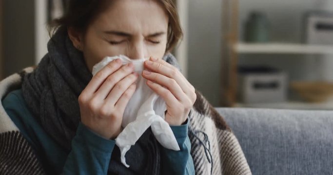 Portrait of the young ill caucasian woman in the warm scarf and covered with a plaid sneezing as she having a cold and fever. In the living room at home. Indoor