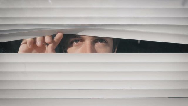 Young man looking out the window through the blinds to the street