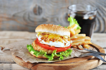 Chickenburger on a round wooden board. Next to the burger is French fries. In the frame, a glass of carbonated beverage and cutlery. Wooden background. Close-up.