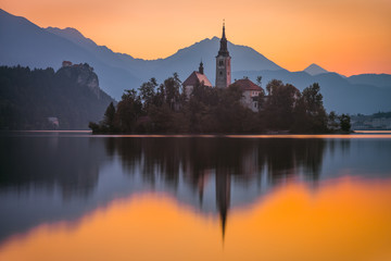 Fototapeta na wymiar Little Island with Catholic Church in Bled Lake, Slovenia at Sunrise with Castle and Mountains in Background