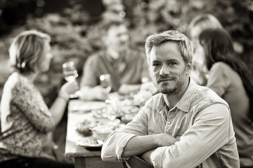 Looking at the camera, a man in his forties sits at a table in the garden with his friends for dinner and have good time