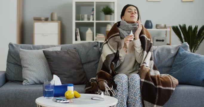 Unhealthy woman in the warm scarf and covered with a plaid sitting on the gray couch in the living room, clogging up her nose with medical drops as she having a flu. Inside