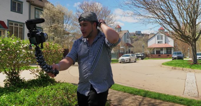 A millennial vlogger wearing a baseball cap talks to his camera while recording a social media video in a residential neighborhood.  	