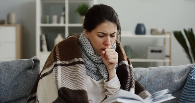 Close up of the young ill caucasian woman in the warm scarf and a plaid reading a book and caughing badly as she having a cold. At home. Inside