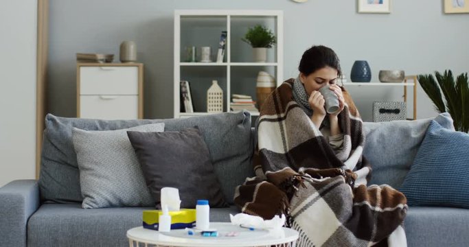 Unwell caucasian young woman sitting on the gray couch in the living room in the warm plaid and drinking hot tea as she having a high temperature during influenza. Indoors