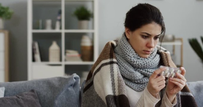 Young sad unwell woman in the warm scarf and covered with a plaid sitting in the cozy room on the sofa, holding a pills and sneezing. Indoor