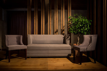 Cinematic Light. A gray sofa and two gray chairs on wooden legs stand on a brown wooden podium, behind the vertical wooden beams and a large wood-pot
