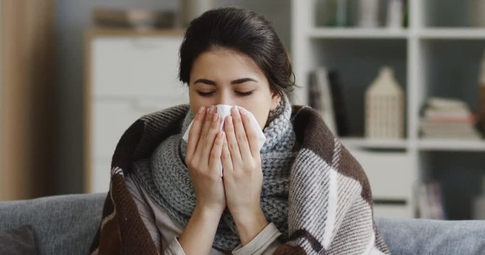 Close up of the young unhealthy caucasian woman in the warm scarf and a plaid blowing her nose in the napkin as she having a cold. At home. Indoors