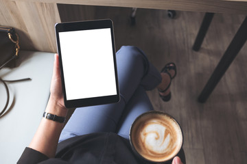 Top view mockup image of a woman sitting cross legged and holding black tablet pc with blank white desktop screen while drinking coffee in cafe