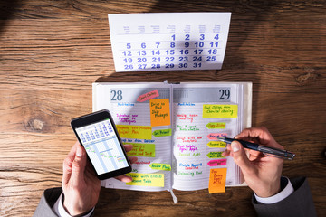 Businessperson With Mobile Phone Writing Schedule In Diary
