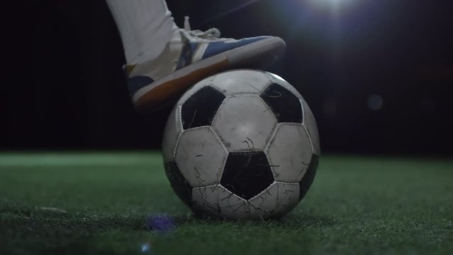 Closeup shot of feet of junior soccer player holding foot on ball on artificial turf in dark arena and then kicking it