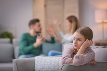 Little unhappy girl sitting in armchair while parents arguing at home