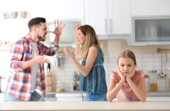 Little Unhappy Girl Sitting At Table While Parents Arguing On Kitchen