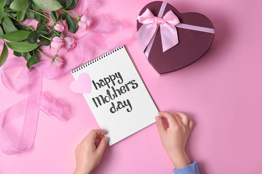 Child holding notebook with phrase "HAPPY MOTHER'S DAY" near gift and roses on table