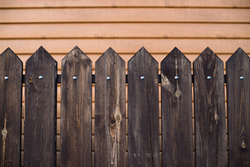 Fototapeta premium Pointed wooden fence on backdrop of house with surface from siding. Background image of wooden fence from rectangular planks with triangular top. Planks fastened crossbar and nails.