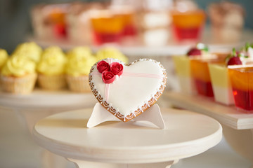 Glazed gingerbread heart stands on the wooden stand in the restaurant. It s decorated with sweet red roses and a little pattern. There is a colorful candybar with cupcakes and jellies behind it.
