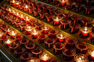 Small white votive candles are lighted and placed in red glass candlesticks among empty ones, lined up on a candle holder stand in a catholic church.
