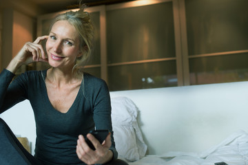 Mature sitting on bed with smartphone in hand, smiling, portrait