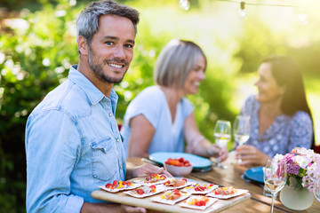  group of friends in their forties gathered around a table in the garden to share a meal. A man offers snacks to guests while looking at the camera