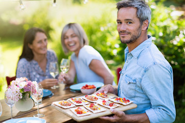  group of friends in their forties gathered around a table in the garden to share a meal. A man offers snacks to guests while looking at the camera