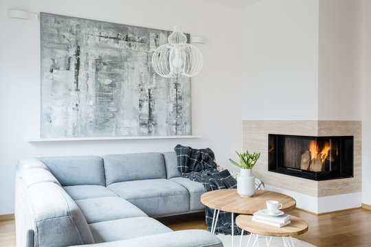 Black blanket thrown on grey corner lounge in white living room interior with fireplace, fresh tulips in vase and big modern painting hanging on the wall