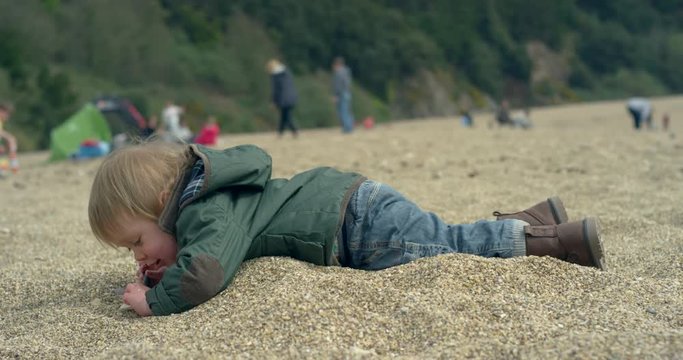 A little toddler boy is lying face down on the beach and is playing with pebbles. Slow motion handheld shot.