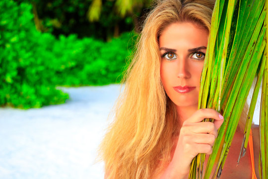 Beautiful blonde woman looks between palm leaves on the sandy beach at the camera