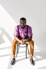 young african american man in stylish clothing with hands in lock sitting on chair on grey background