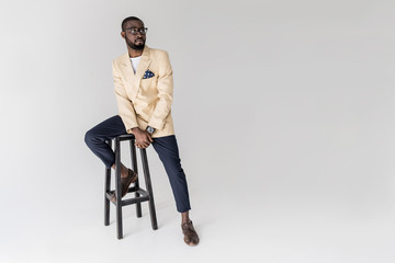 full length view of stylish african american man in eyeglasses sitting on stool and looking away...