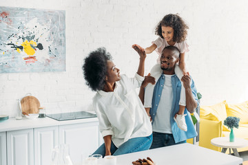 african american daughter sitting on fathers shoulders and giving high five to mother in kitchen at home - Powered by Adobe