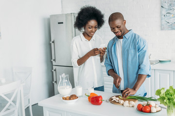 Fototapeta na wymiar smiling african american man cooking breakfast and wife with smartphone standing near by in kitchen at home