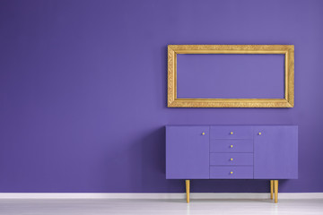 Real photo of a golden frame hanging above a purple cupboard in modern and simple living room...