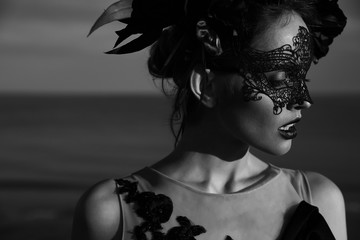 Black and white dramatic portrait of beautiful young woman in dress with black flowers and with black lace mask on face and floral crown on head. Sea background - 202467541