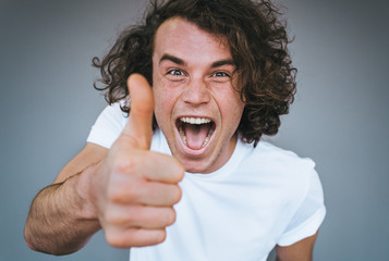 Closeup shot of handsome male smling with healthy toothy smile, wears white t-shirt looking at the camera with positive expression, showing thumbs up posing on grey wall.Body language, emotion concept