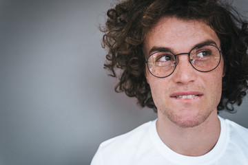 Obraz premium Pensive handsome pensive male with curly hair, wears white t-shirt and trendy round spectacles, looking up aside and thinking about ideas, bites his lips isolated on grey wall. People concept.