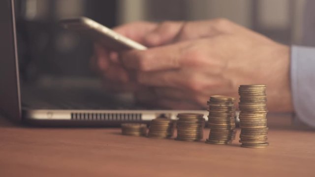 Businessman using smartphone with stacked coins in foreground, close up shot with selective focus