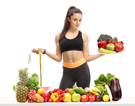 Fitness girl holding a plate and a measuring tape behind a table with fruit and vegetables