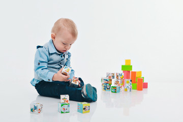 blond boy in blue suit plays with colorful blocks on white background. A small child builds a tower of cubes on the floor