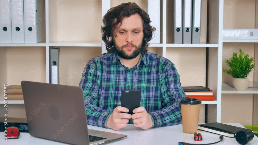 Poster engineer sitting at the workspace using smartphone texting message and drinking coffee. handsome bea