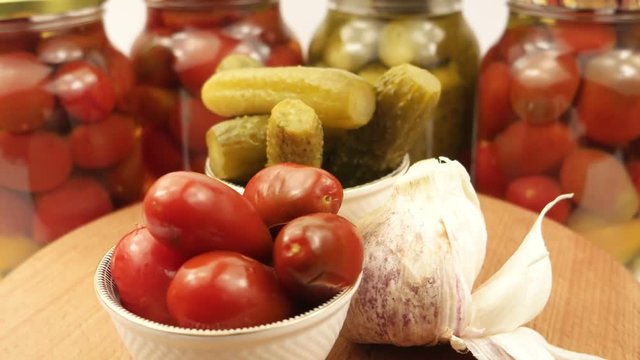 Two small bowls of the pickled tomatoes and cucumbers, garlic head on the background of glass cans with tomatoes and cucumbers