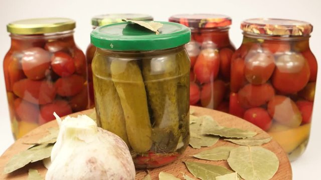 Glass can of canned cucumbers and garlic head in the background of cans with tomatoes