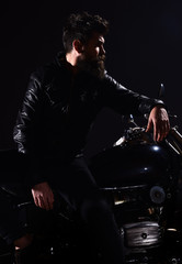 Man with beard, biker in leather jacket lean on motor bike in darkness, black background. Macho, brutal biker in leather jacket stand near motorcycle at night time. Brutality and masculine concept.