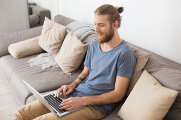 Portrait of young man sitting on big gray sofa and working on laptop at home isolated