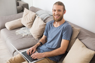 Fototapeta premium Portrait of young smiling man sitting on big gray sofa with laptop and happily looking in camera isolated